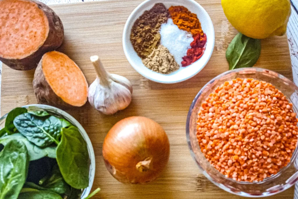 A wooden tray displaying soup ingredients of red lentils, spinach, onion, garlic, lemon and spices.